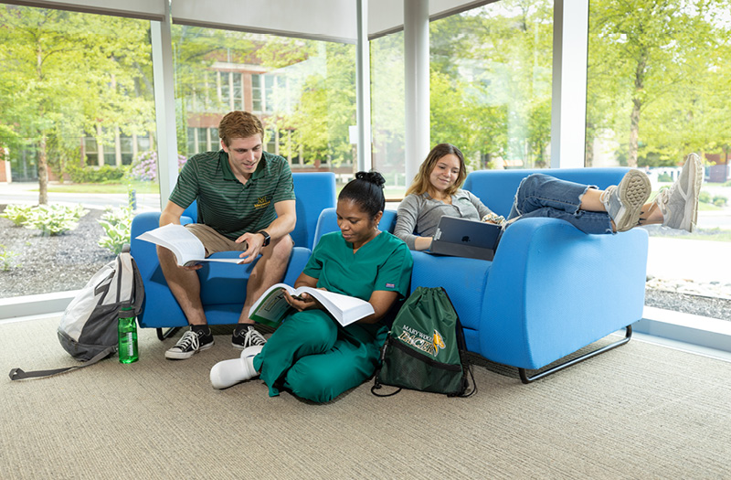 Three students sitting comfortably on oversized furniture in a library setting.